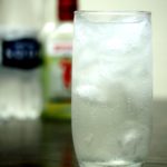 Un grand verre de Tom Collins avec une bouteille de soda water et de gin en arrière plan
